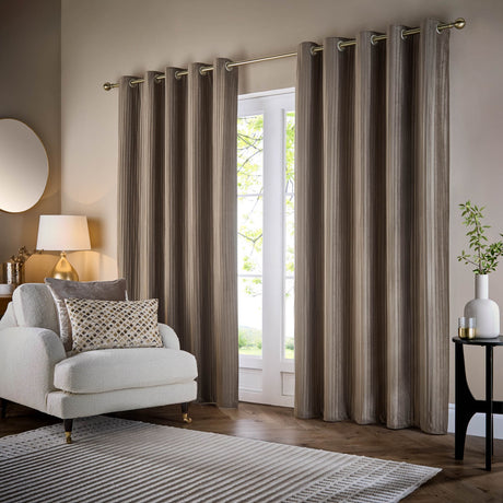 Corinthian velvet lined eyelet curtains in taupe, hung on a brass rail beside a window in a living room, showing fine vertical micro-pleat texture in warm greyish-brown velvet.