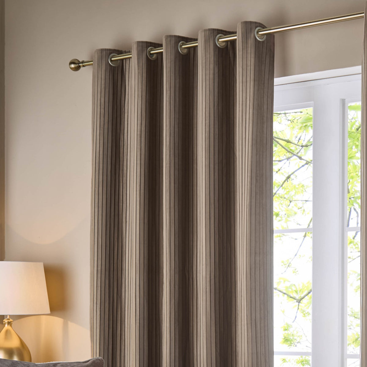 Corinthian Velvet Lined Eyelet Curtains in taupe with brass eyelets on a metal pole, showing fine vertical micro-pleat texture in a living room beside a window.