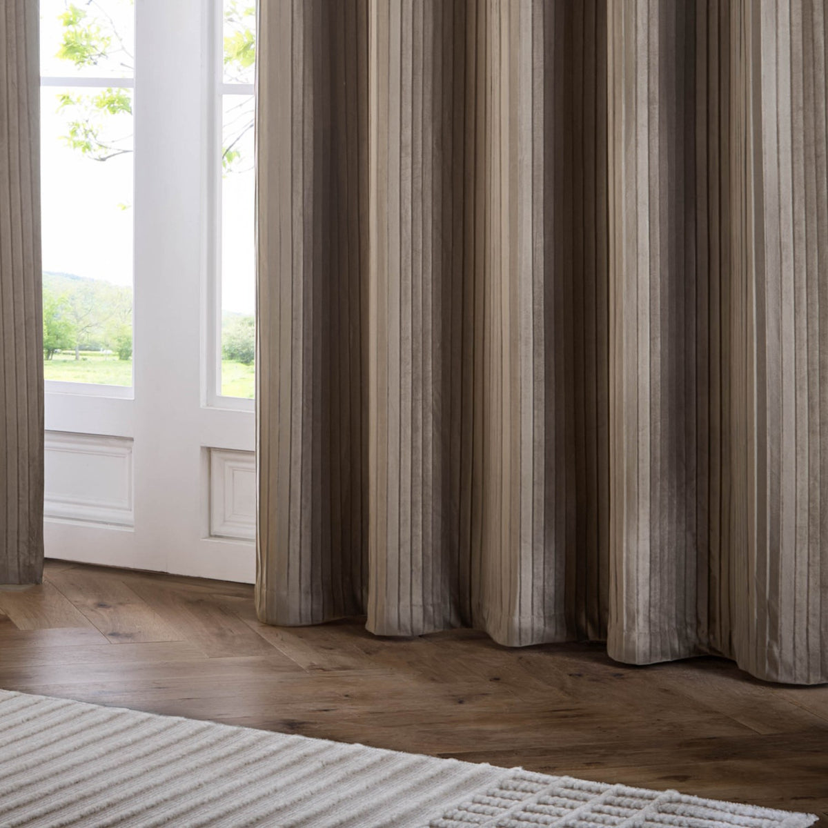 Corinthian Velvet Lined Eyelet Curtains in taupe, showing fine micro-pleated velvet fabric pooling on a wooden floor beside a glazed door in a living room setting.