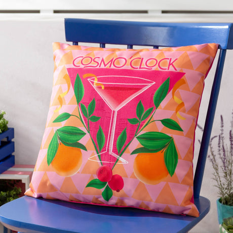 Cosmo O' Clock square polyester cushion in pink, printed with a cocktail glass, oranges, leaves and berries, shown on a blue chair.