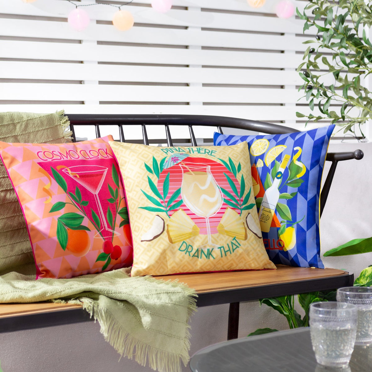 Cosmo O' Clock square cushion in pink polyester with cocktail glass print, oranges and berries, styled on an outdoor bench beside coordinating printed cushions.