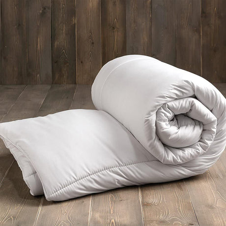 Cosy Home Anti-Allergy Hollowfibre Quilt shown rolled and partly unfolded in white polyester-cotton casing on a wooden floor against a dark wood panel background.