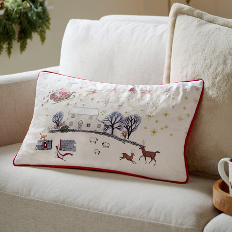 Cotswold Christmas Embroidered Velvet Cushion in cream, rectangular with red piped trim, showing a festive village scene with Santa, reindeer and sheep, styled on a sofa.