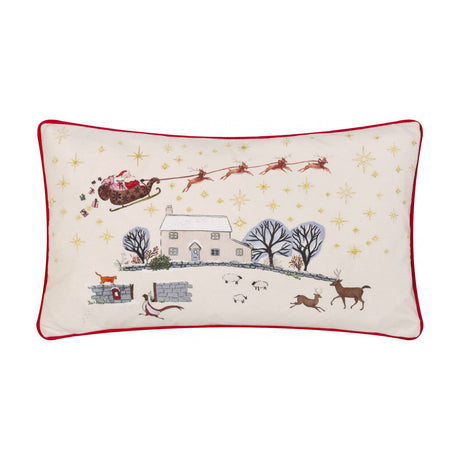 Rectangular Cotswold Christmas embroidered velvet cushion in cream, depicting Santa's sleigh, reindeer, a snowy farmhouse and sheep, edged with red piped trim, shown on a white background.