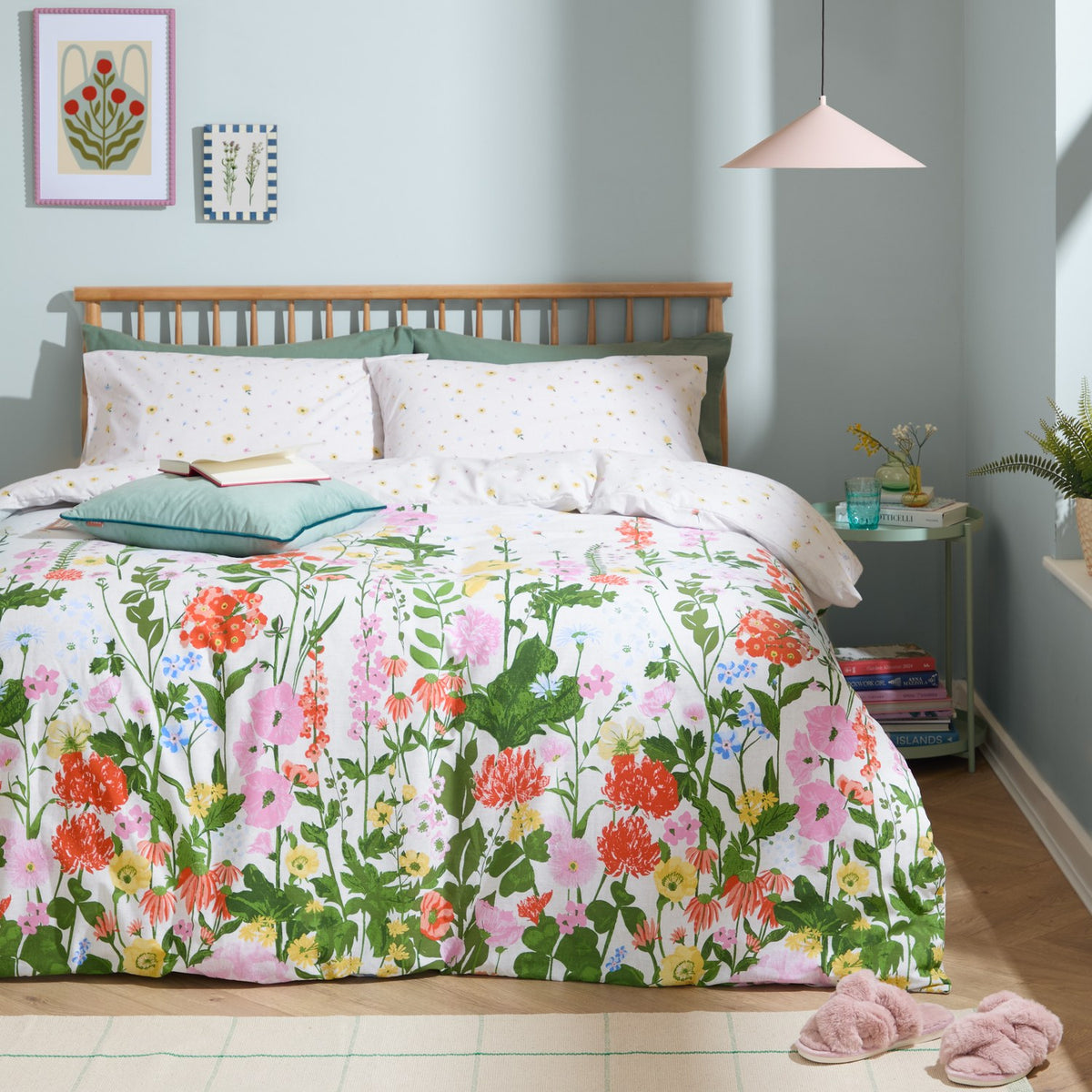 Cottage Garden reversible duvet cover set on a bed, with a multicoloured floral border print in pink, coral, yellow and green, styled with ditsy-print pillowcases in a pale bedroom setting.