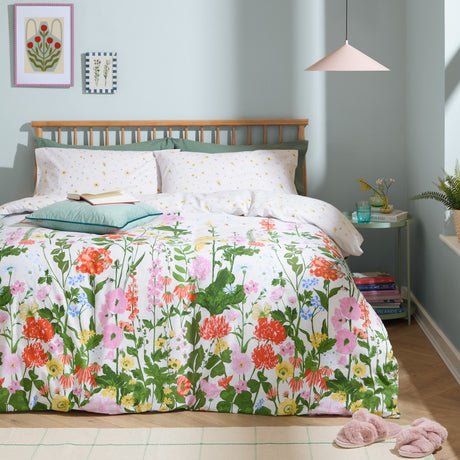 Cottage Garden reversible duvet cover set on a bed, with a multicoloured floral border print in pink, coral, yellow and green, styled with ditsy-print pillowcases in a pale bedroom setting.