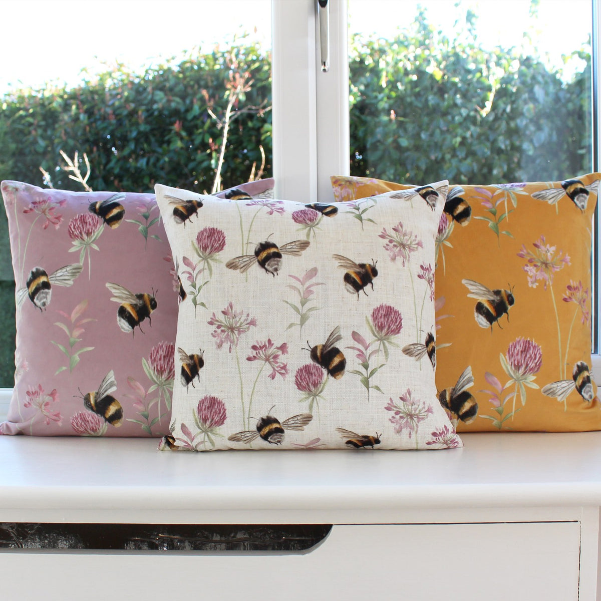 Country Animal Velvet Cushion in heather pink with bumblebee and clover print, shown on a windowsill beside cream and mustard bee-print cushions.
