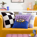 Cozee Check faux fur cushion in black and white checkerboard, shown on a mustard sofa alongside patterned and eye-print cushions in a living room setting.
