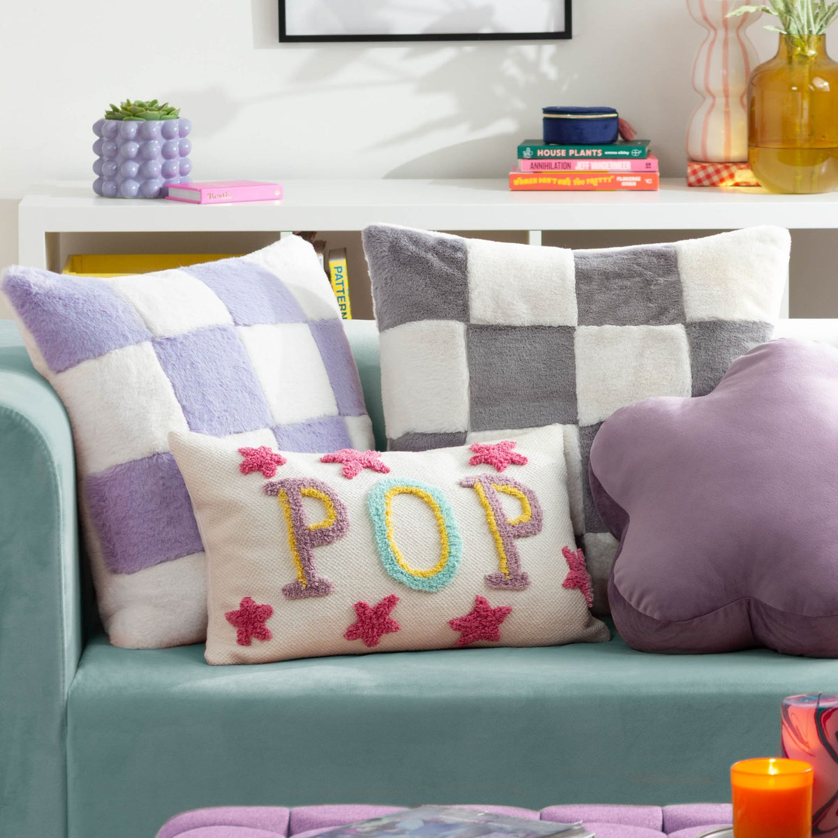 Cozee Check Plain Faux Fur Cushion in lilac and white checkerboard, 50 x 50cm, shown on a teal sofa alongside patterned cushions in a living room setting.