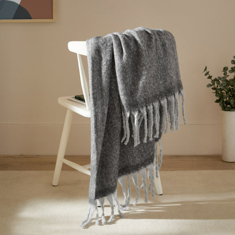Cosy Plain Polyester Throw in charcoal grey with white tasselled fringe, draped over a white wooden chair in a living room setting.