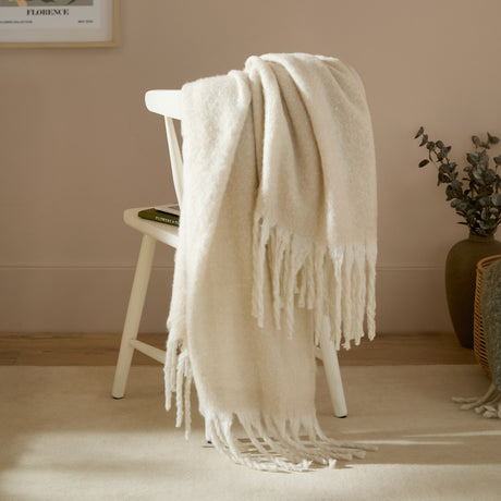 Cosy Plain Polyester Throw in ecru draped over a white wooden chair, showing its brushed texture and knotted fringe in a warm living room setting.