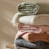 Cosy Plain Polyester Throw in ecru, folded with knotted fringe, shown in a stack of neutral and pastel throws in green, pink, rust, and grey.