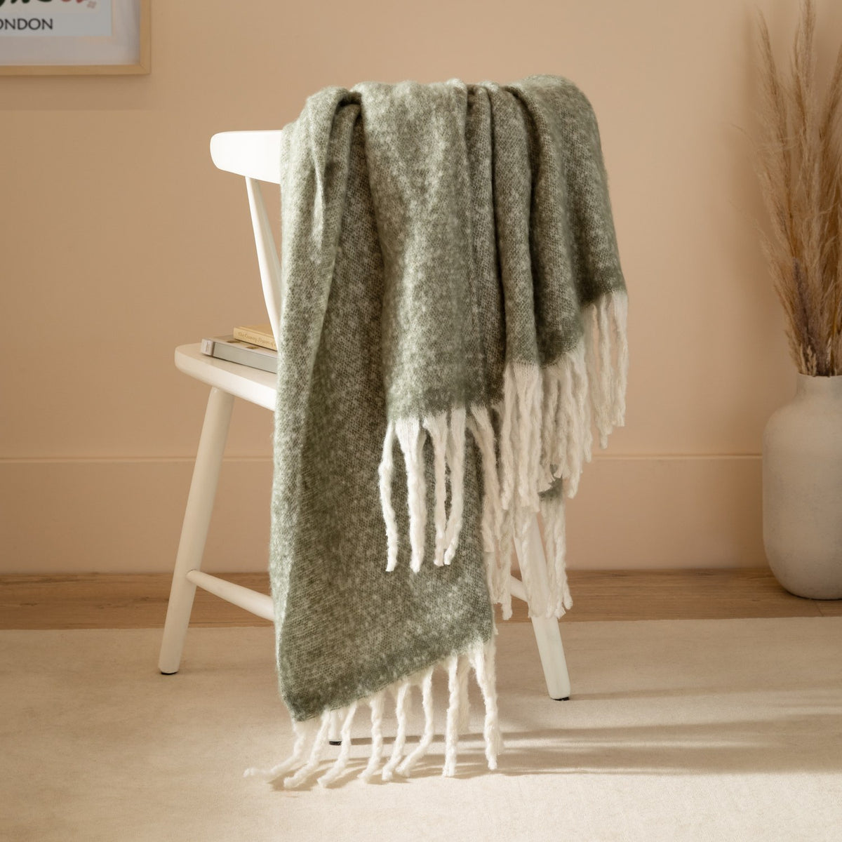 Cosy Plain Polyester Throw in Green draped over a white chair, showing its woven sage-toned body and chunky ivory fringed trim in a warm-toned living room setting.