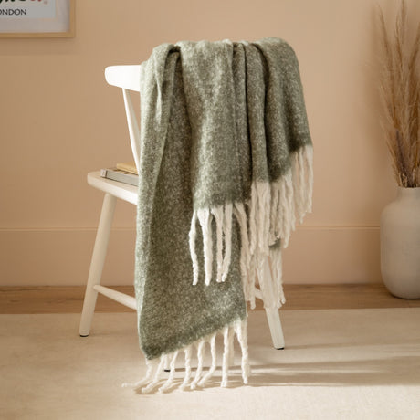 Cosy Plain Polyester Throw in Green draped over a white chair, showing its woven sage-toned body and chunky ivory fringed trim in a warm-toned living room setting.
