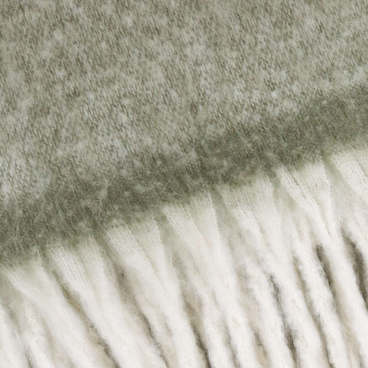 Close-up of the Cosy Plain Polyester Throw in green, showing the woven sage-grey body meeting a chunky ivory fringed trim.