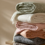 Cosy Plain Polyester Throw in pink, folded with brushed texture, shown stacked with green, cream, orange, and grey throws against a neutral background.