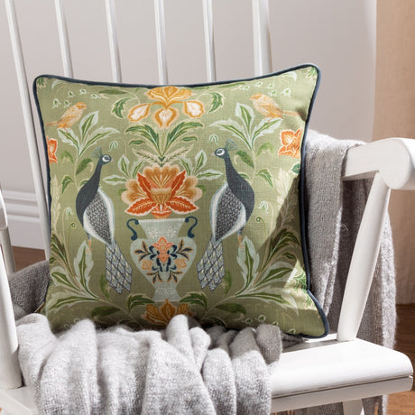 Chatsworth Peacock cushion in sage green linen, 43 x 43cm, with a symmetrical peacock and floral urn design, navy piped edge, shown on a white armchair with a grey knit throw.