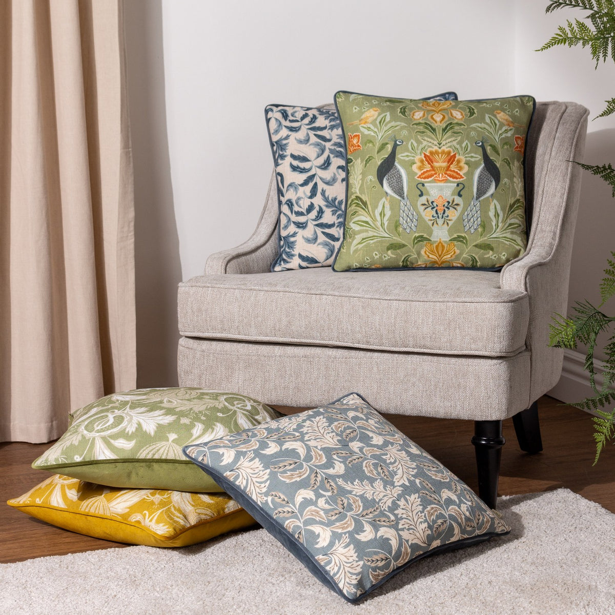 Chatsworth Peacock Velvet Linen Cushion in sage, styled on a beige armchair with coordinating floral and blue cushions in a living room setting.