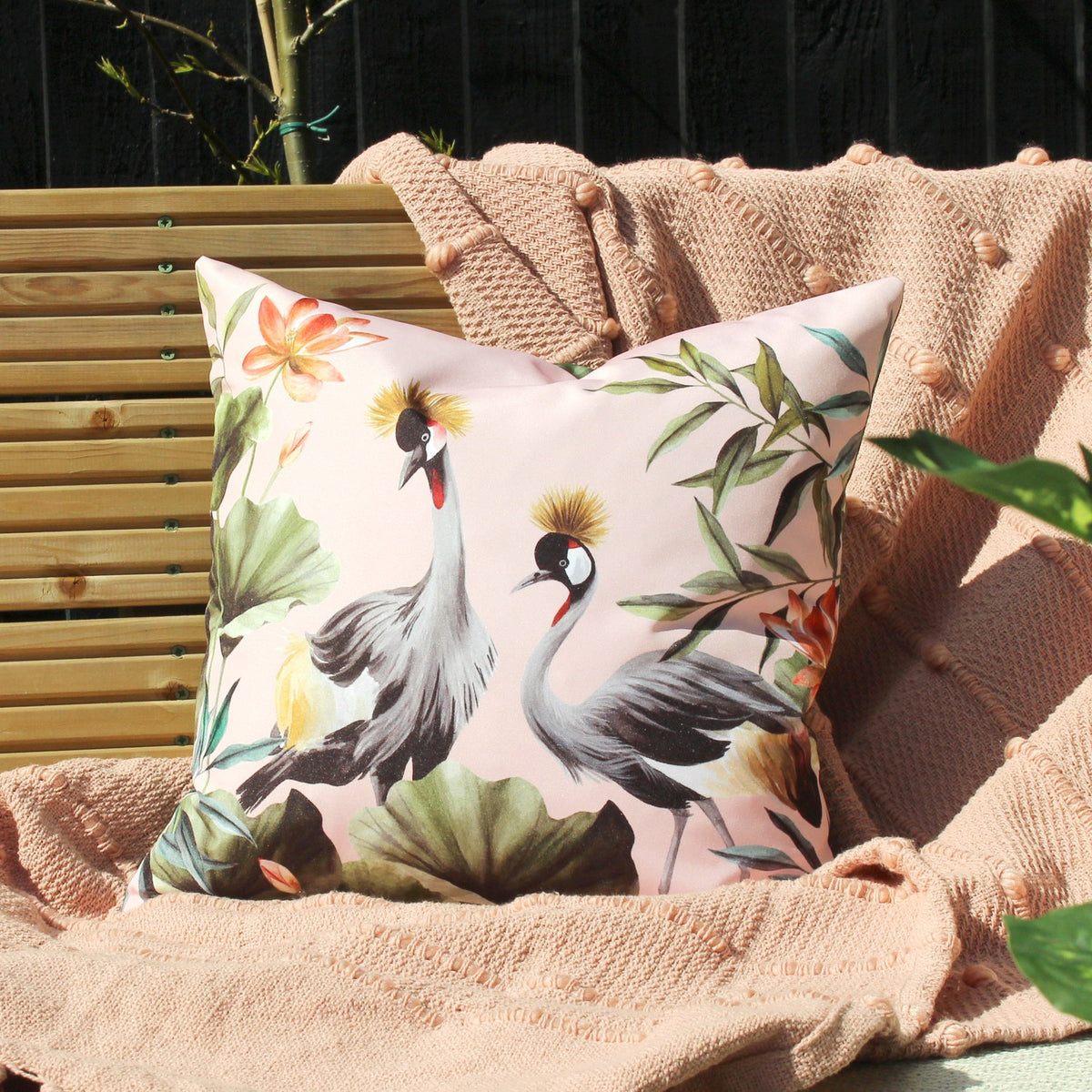 Square blush cushion with crowned crane birds, tropical leaves and lotus flowers, shown on a pink knitted throw against a wooden bench.