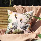 Square blush cushion with crowned crane birds, tropical leaves and lotus flowers, shown on a pink knitted throw against a wooden bench.