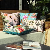 Cranes Reversible Polyester Cushion in blush with crowned crane and tropical leaf print, styled among patterned cushions on an outdoor bench with plants.