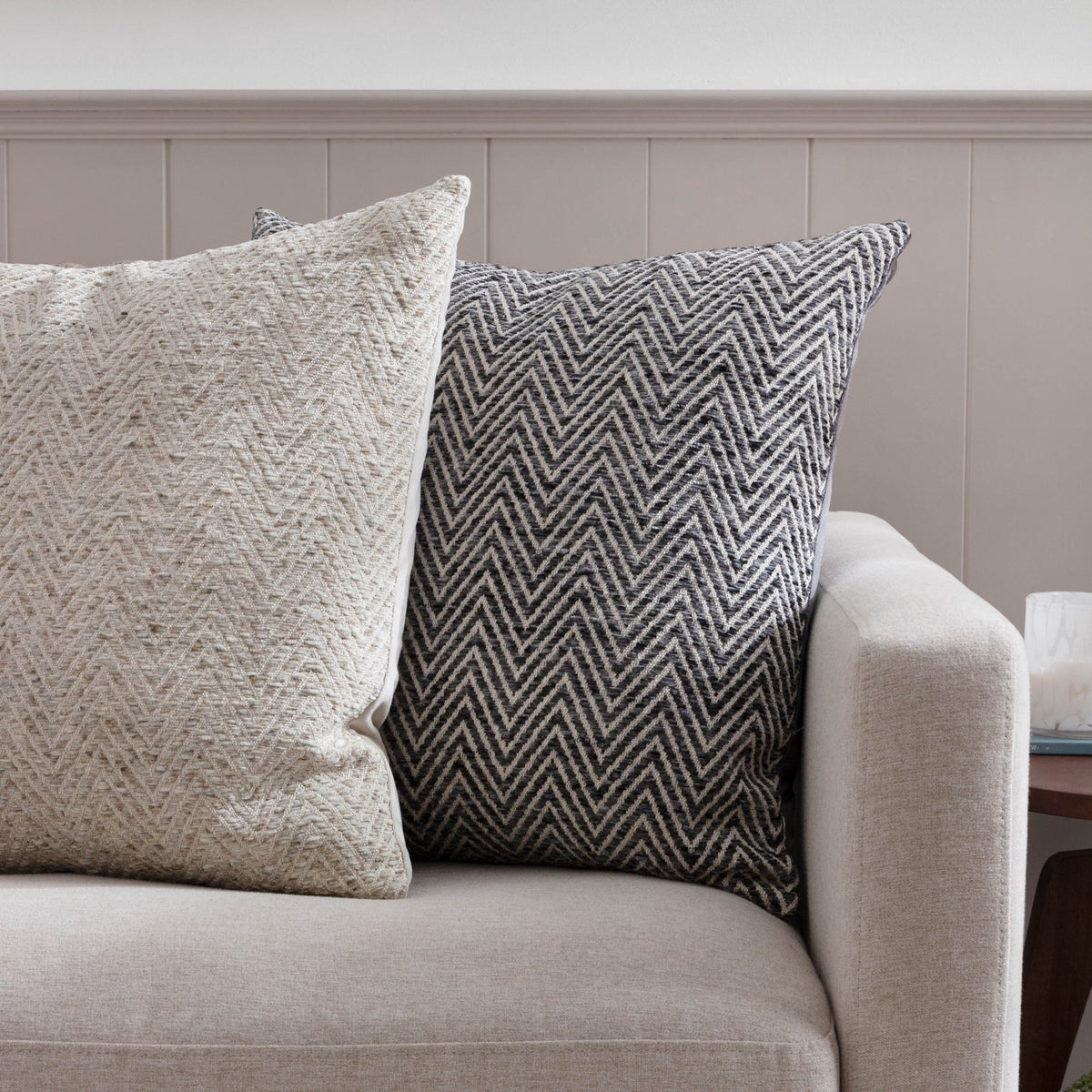Croft Plain Velvet Cushion in charcoal, square with a woven chevron front in charcoal and cream, shown on a beige sofa beside a cream herringbone cushion.