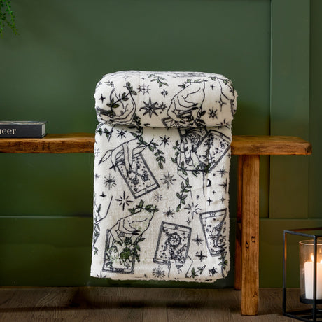 Celestial Stories Printed Polyester Throw in cream draped over a wooden bench, showing tarot card, star and trailing leaf line-work with green foliage detailing.