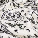 Celestial Stories printed polyester throw in cream, shown draped in folds with charcoal tarot card, hand, star, moon and green foliage motifs.