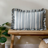 Carmon Stripe Plain Linen Cushion in blue and cream vertical stripes with a gathered ruffle trim edge, shown on a wooden bench in a living room setting.