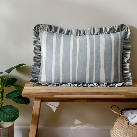Carmon Stripe Plain Linen Cushion in blue and cream vertical stripes with a gathered ruffle trim edge, shown on a wooden bench in a living room setting.