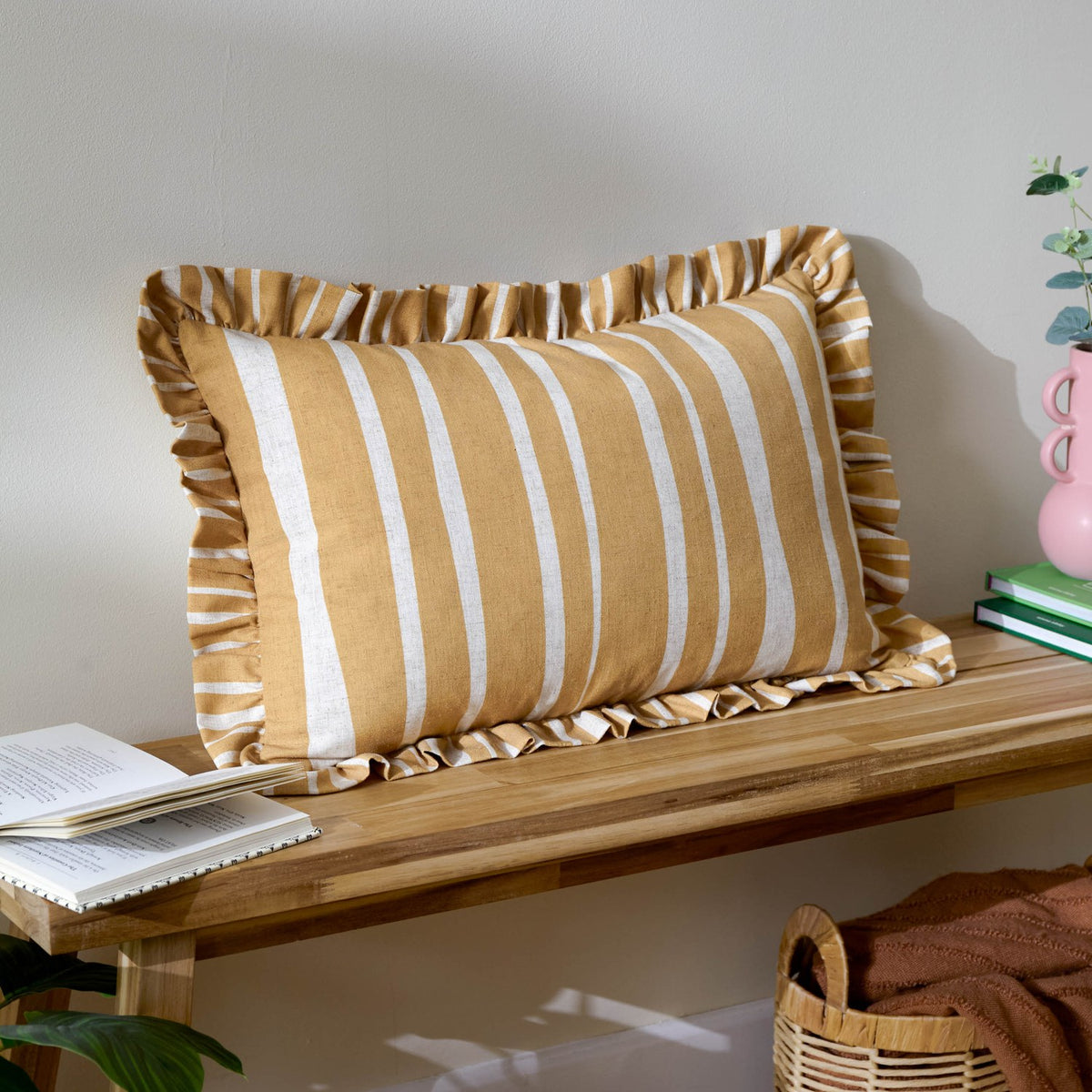 Carmon Stripe rectangular linen cushion in ochre with cream vertical stripes and a gathered ruffle trim, shown on a wooden bench beside books and a plant.
