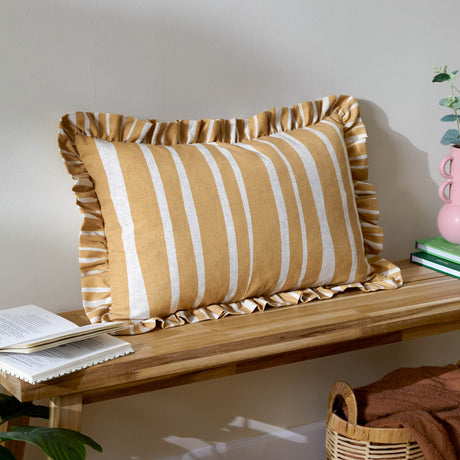 Carmon Stripe rectangular linen cushion in ochre with cream vertical stripes and a gathered ruffle trim, shown on a wooden bench beside books and a plant.