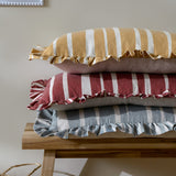 Carmon Stripe Plain Linen Cushion in ochre with cream stripes and ruffle trim, stacked with red and blue striped cushions on a wooden bench.