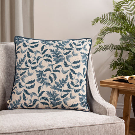 Chatsworth Topiary Velvet Linen Cushion in Petrol, showing the printed leaf-patterned linen front with petrol blue piping, styled on a grey armchair beside a plant.