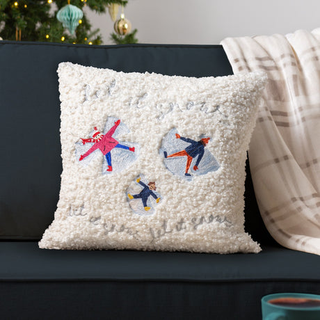 Cream boucle cushion with embroidered snow angel figures and "let it snow" script, styled on a dark sofa beside a Christmas tree.