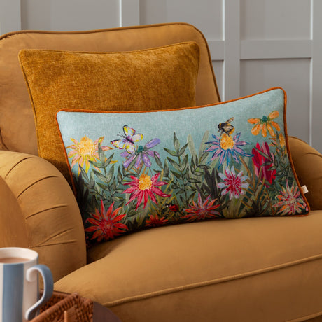 Country Wilds Blossom Meadow rectangular cushion in sky blue with rust piping, featuring a painted floral meadow, butterfly, bee and ladybird, shown on a mustard armchair.