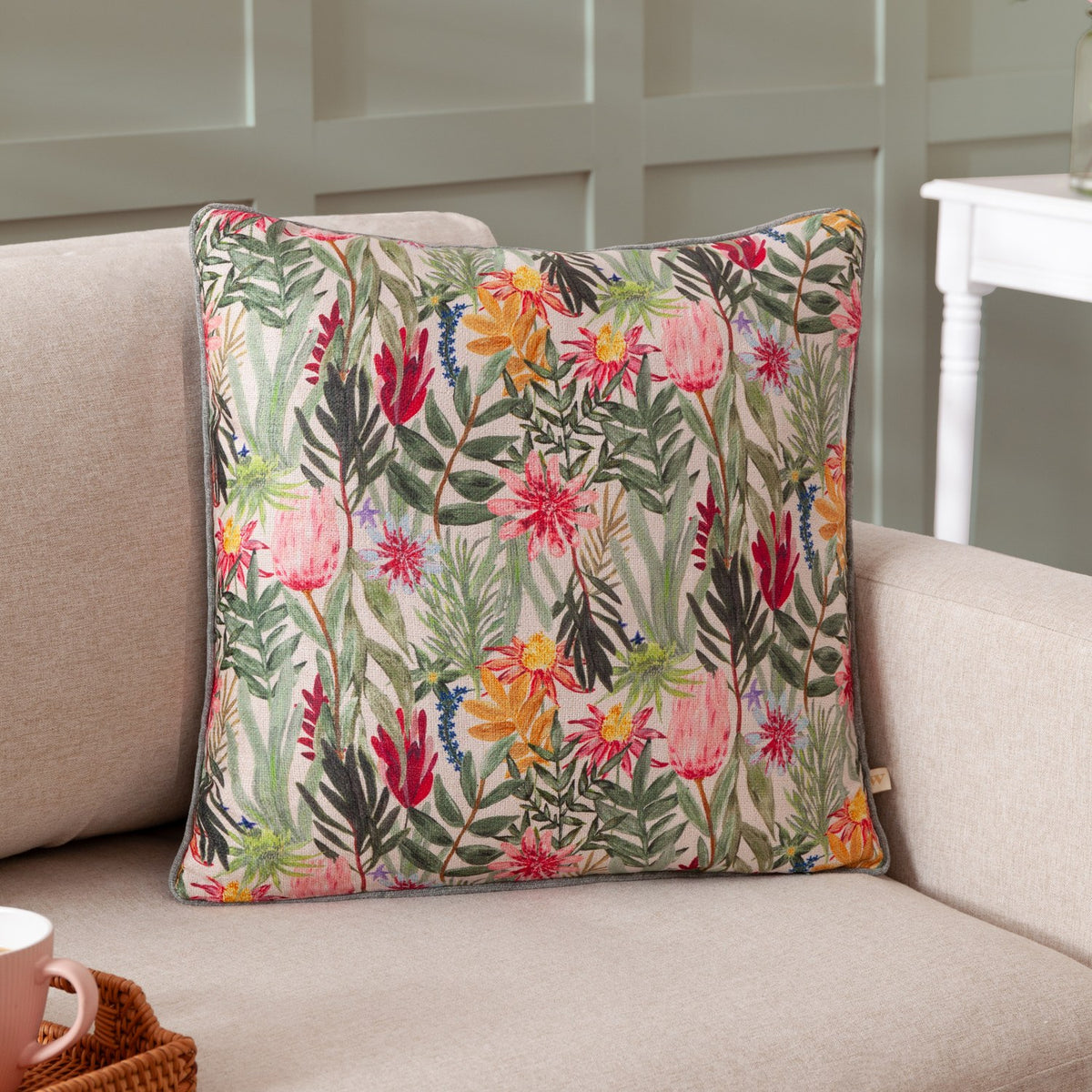 Country Wilds Blossom Floral polyester cushion in cream with pink, yellow and red florals and green foliage, piped edge, shown on a beige sofa.