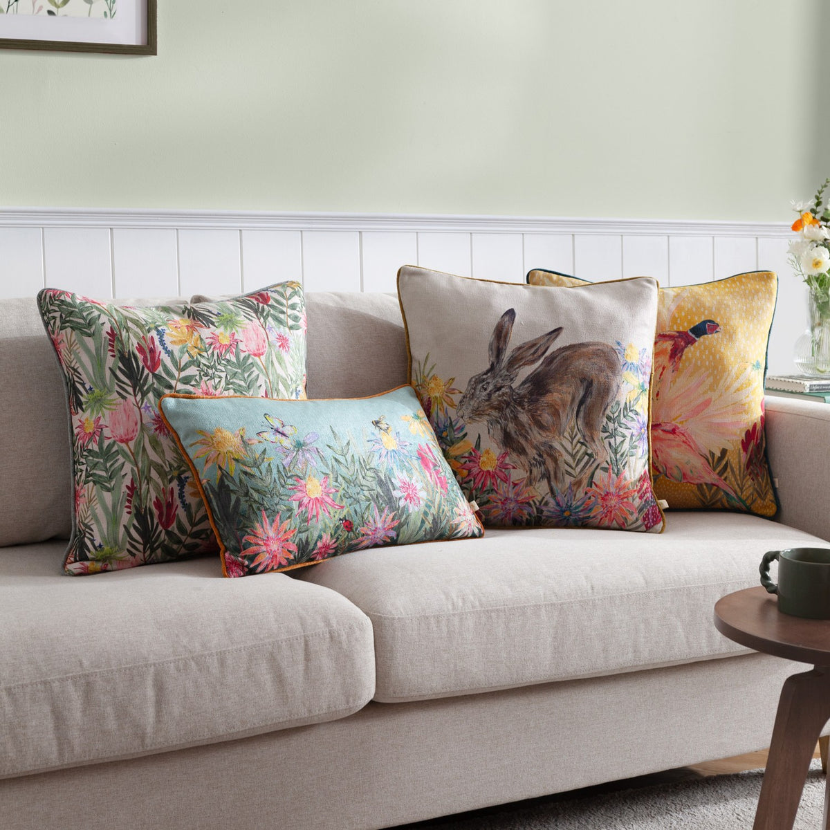 Country Wilds Blossom Floral polyester cushion in cream with pink, mustard and red florals, shown among patterned cushions on a beige sofa in a living room.