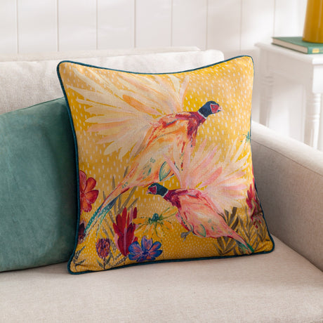 Country Wilds Pheasant Bloom polyester cushion in gold, showing pheasants and wildflowers, teal piping, styled on a beige sofa with a teal cushion.