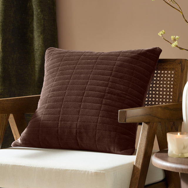 Dales quilted velvet cotton cushion in brown with a square shape, shown on a wooden armchair in a living room setting.