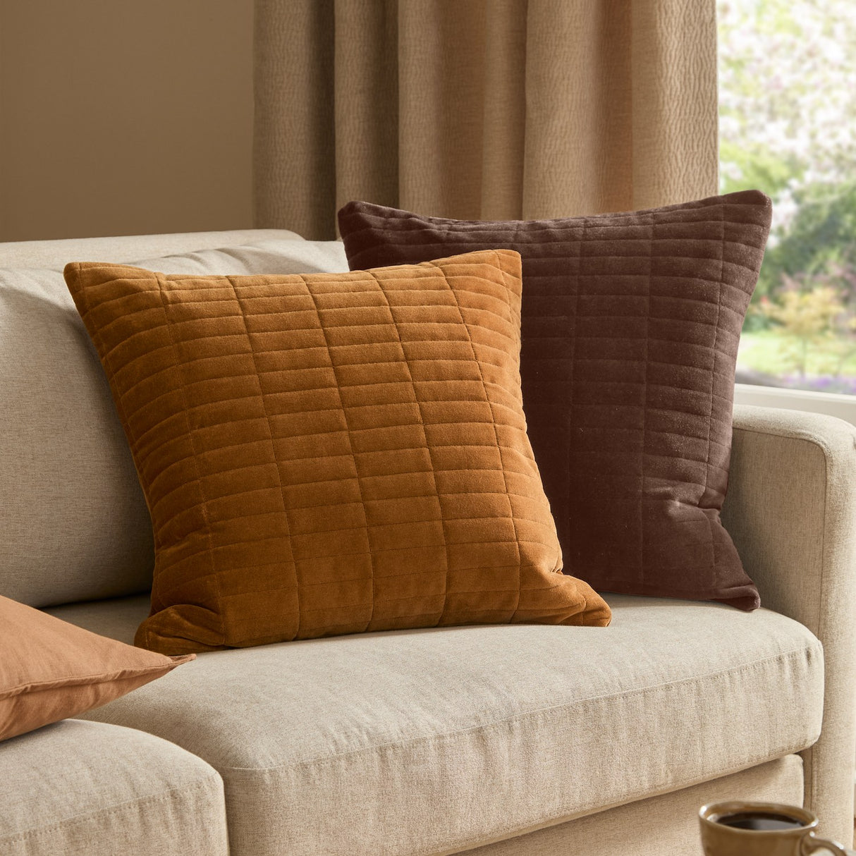 Dales quilted velvet cotton cushion in brown with grid-stitched texture, shown on a beige sofa beside a matching tan cushion.