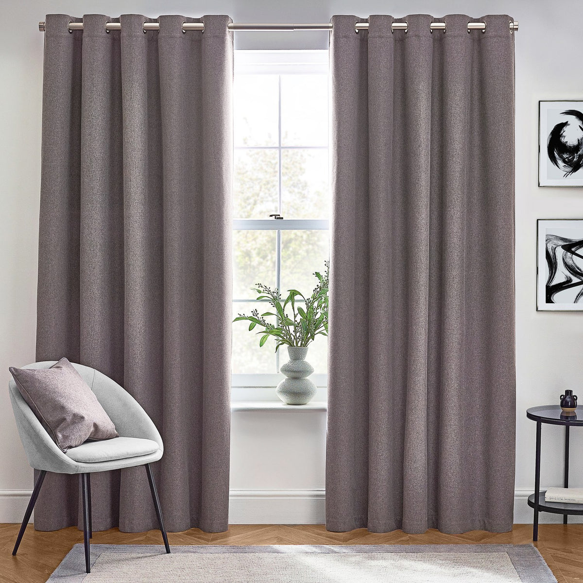 Dawn Plain Reversible Eyelet Curtains in charcoal grey hung floor-length on a metal pole in a living room, showing the fabric's textured finish beside a window seating area.
