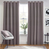 Dawn Plain Reversible Eyelet Curtains in charcoal grey hung floor-length on a metal pole in a living room, showing the fabric's textured finish beside a window seating area.