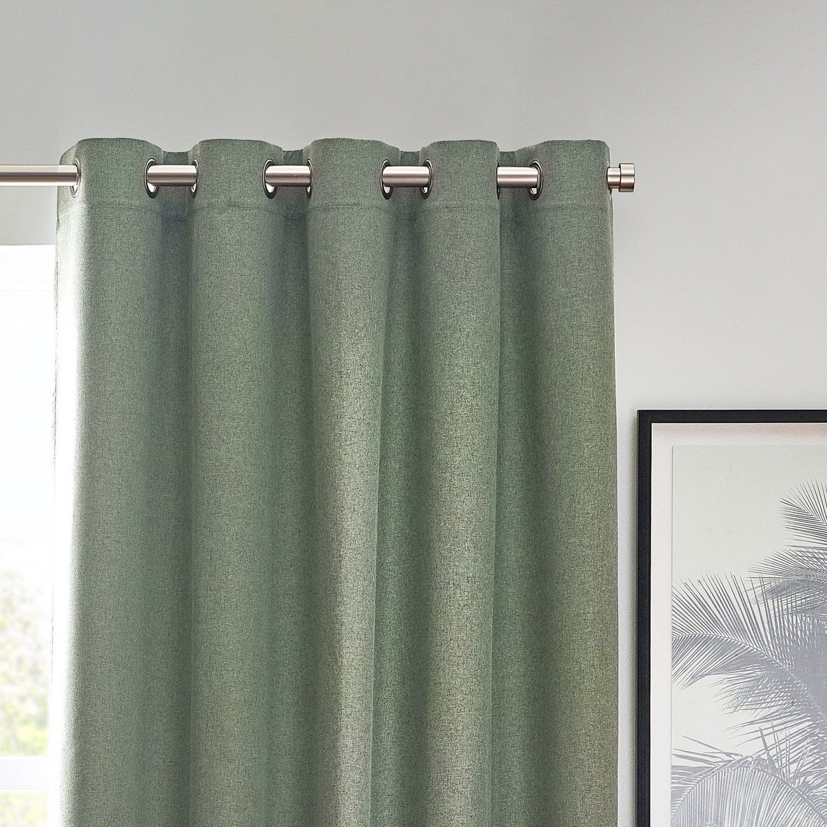 Dawn Plain Reversible Eyelet Curtains in Eucalyptus, dusty green marl-effect fabric with silver eyelet grommets on a metal pole, shown against a light wall near a framed print.