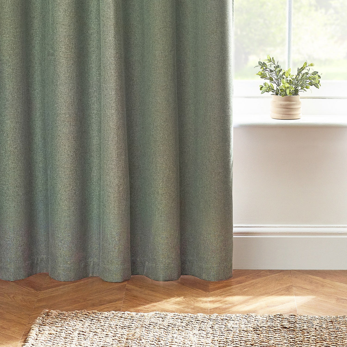 Dawn Plain Reversible Eyelet Curtains in Eucalyptus, dusty green marl-effect fabric with eyelet header, styled in a living room beside a window with a potted plant on the sill.