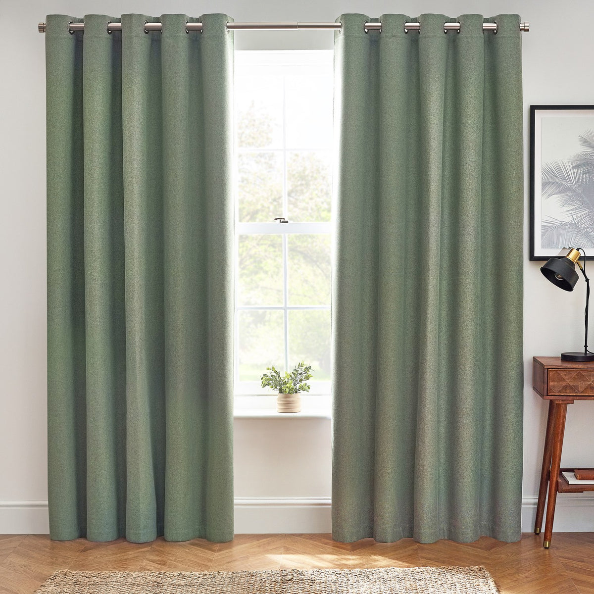 Dawn Plain Reversible Eyelet Curtains in eucalyptus green, floor-length with metal eyelet heading on a pole, shown in a living room beside a window and wooden side table.
