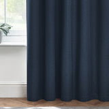 Dawn Plain Lined Eyelet Curtain in midnight blue polyester fabric, shown hanging in soft folds beside a window with a plant on the sill.