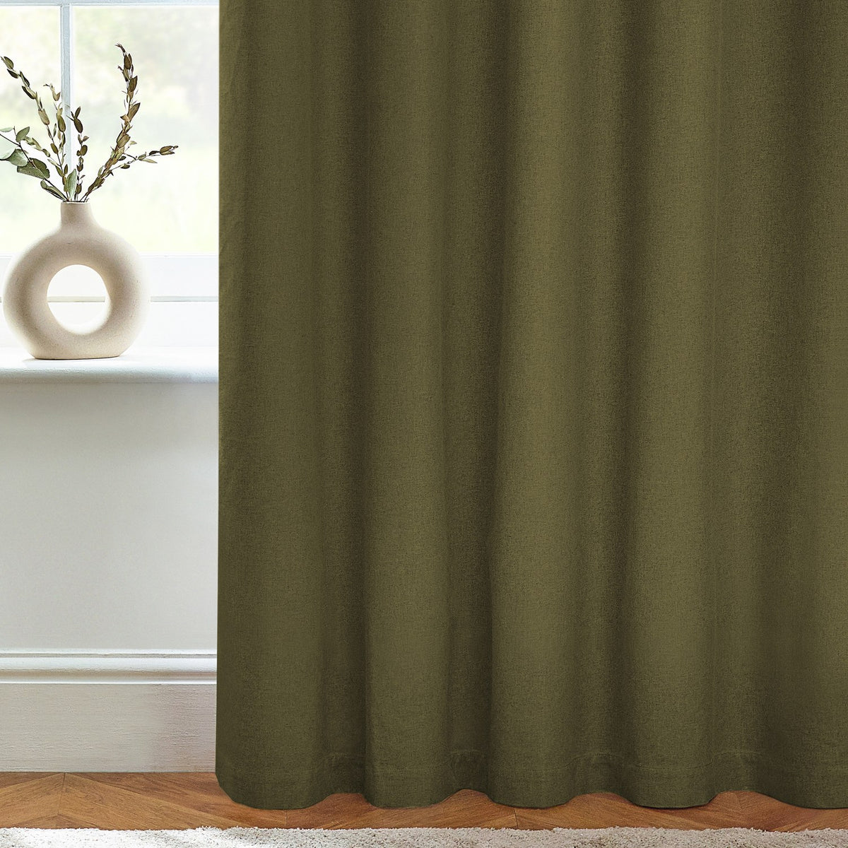 Dawn Plain Lined Eyelet Curtains in olive green fabric with soft textured weave, shown hanging in a soft furnishings arrangement beside a window with a cream vase on the sill.