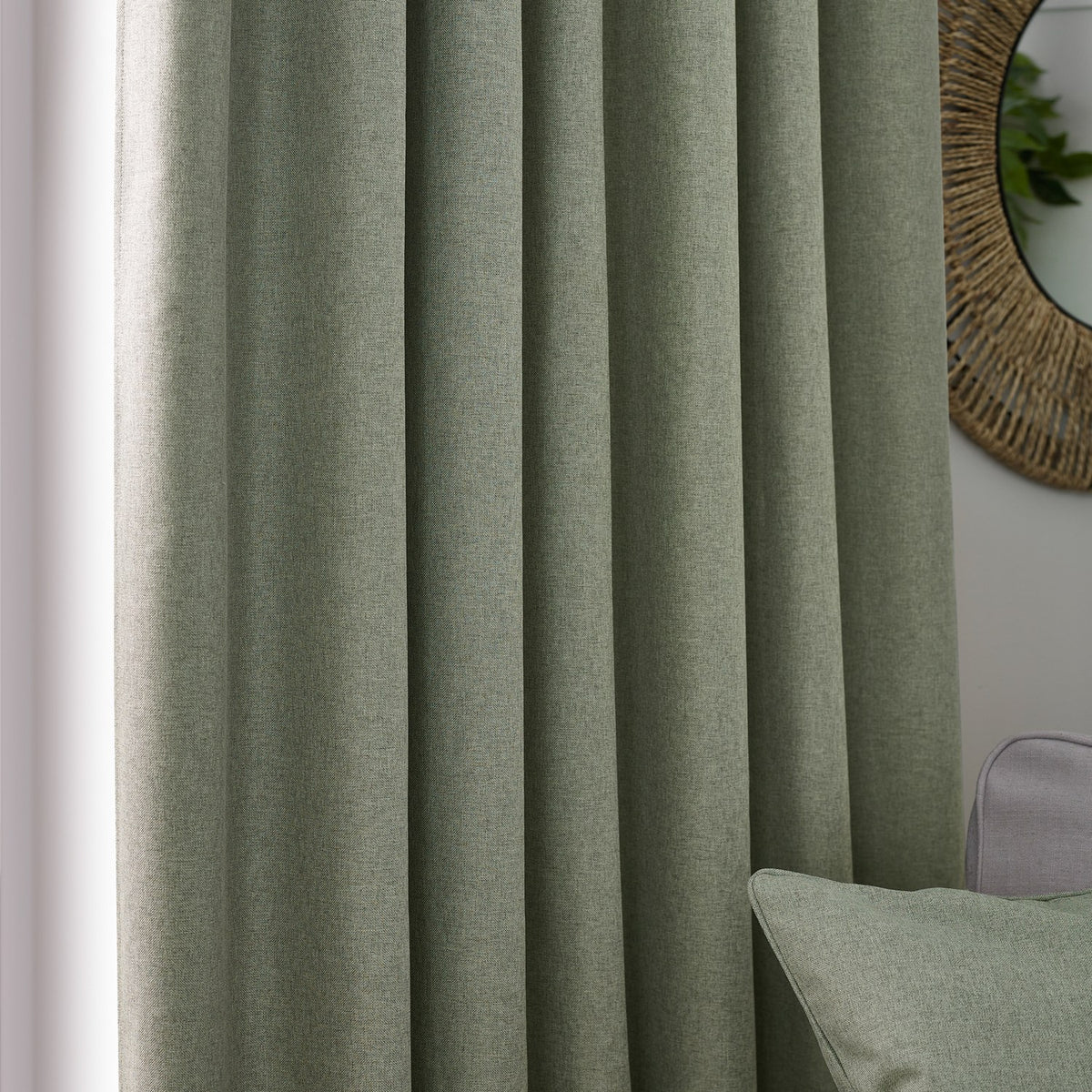 Dawn Reversible Pencil Pleat Curtains in eucalyptus green polyester with soft marl texture, shown pleated in a living room beside a rattan mirror and matching cushion.