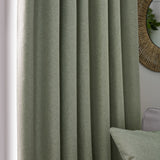 Dawn Reversible Pencil Pleat Curtains in eucalyptus green polyester with soft marl texture, shown pleated in a living room beside a rattan mirror and matching cushion.
